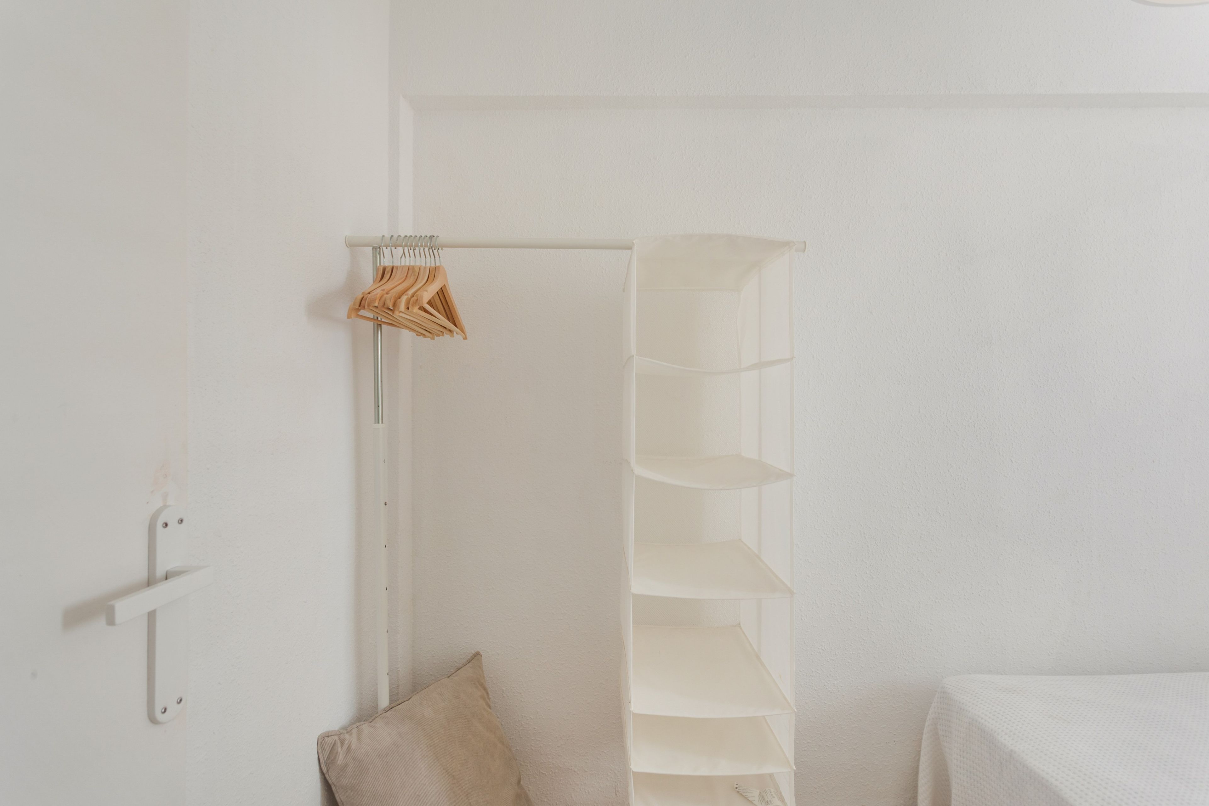 Quarto branco com um cabideiro de madeira, uma estante branca e uma almofada bege.