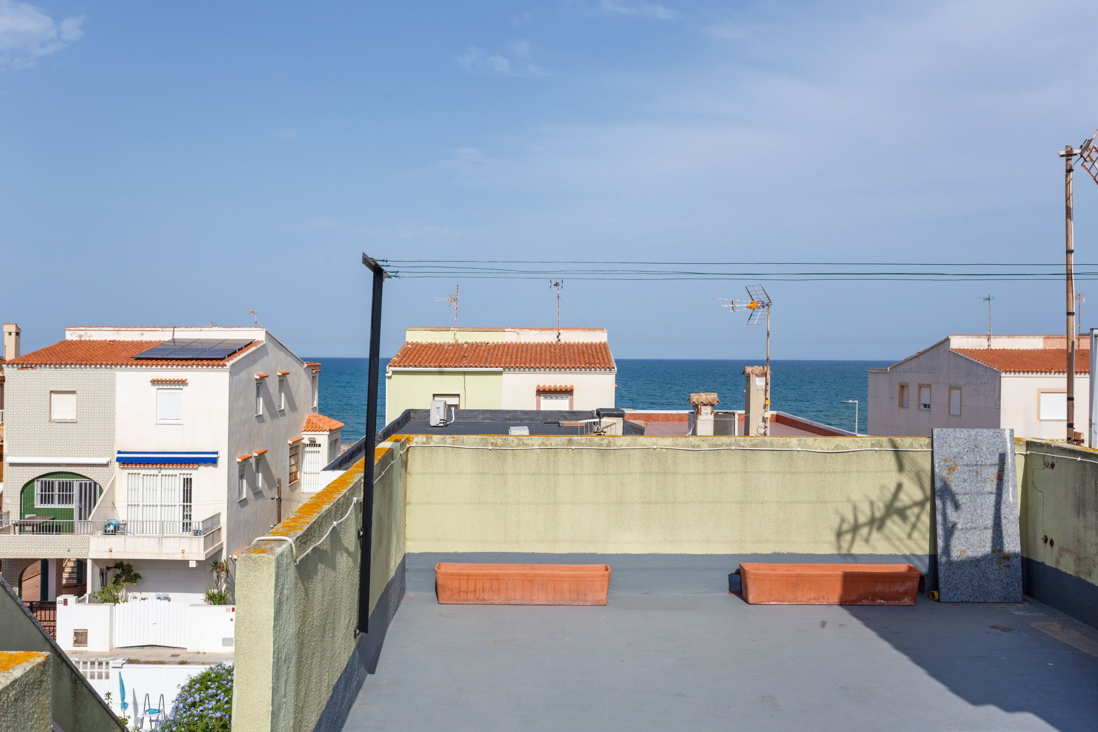 A rooftop terrace with a view of the sea, featuring a concrete floor, two wooden benches, and a metal railing.