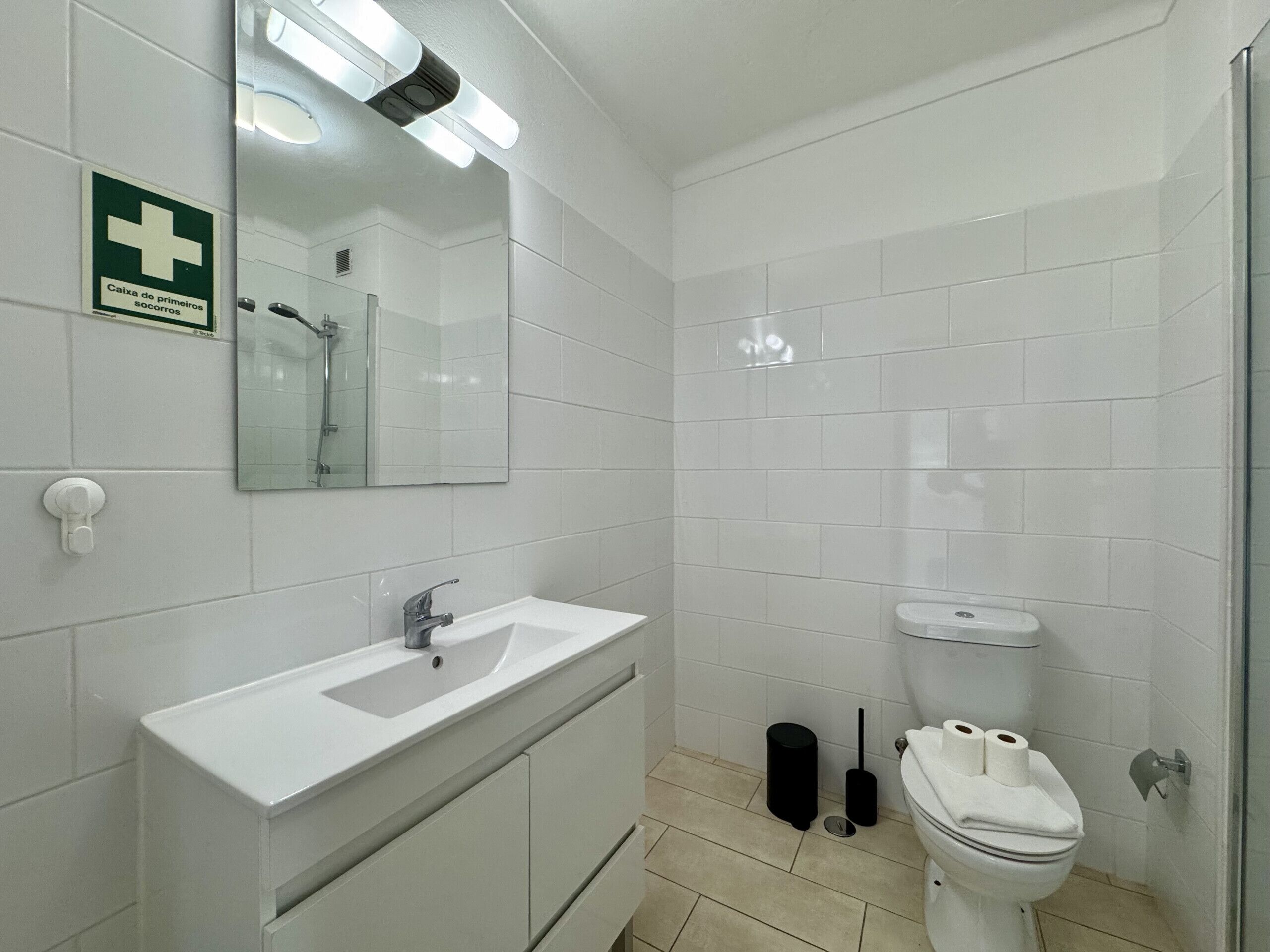 Un baño con lavamanos blanco, espejo y retrete. Hay un letrero sobre el lavamanos y un cesto de basura junto al inodoro.