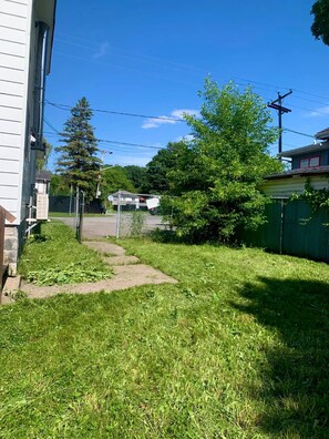 A grassy yard with a pathway leading to a house, surrounded by trees and a fence.