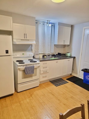 A compact kitchen with a stove, oven, and refrigerator. There is a sink with a faucet, a countertop, and cabinets. A trash bin is visible.