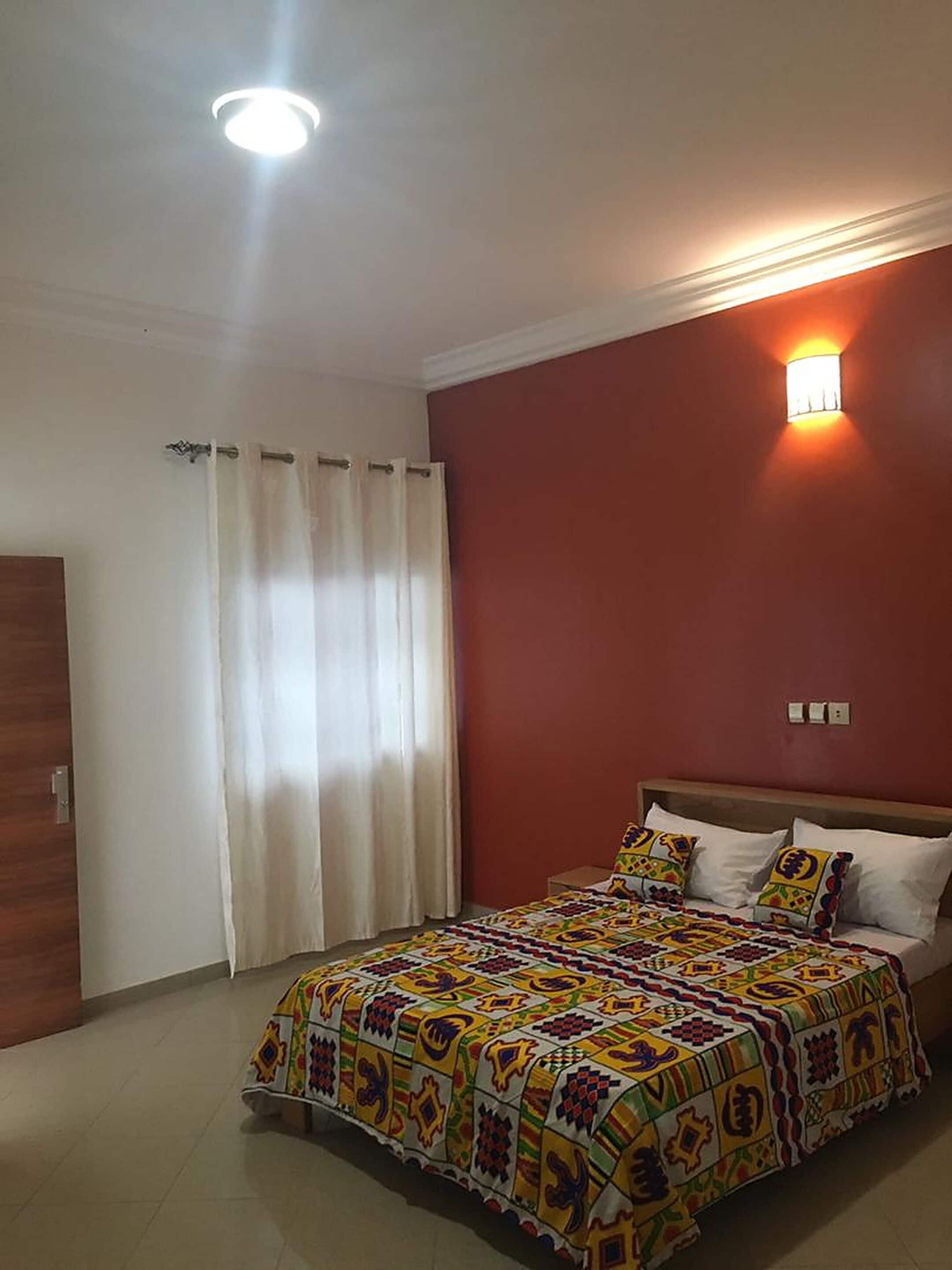 Une chambre à coucher avec un lit, des rideaux, une lampe fixée au mur et un luminaire au plafond.