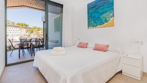 Une chambre d’hôtel avec un lit, un tableau représentant une plage, un bureau avec une lampe et une vue sur l’extérieur.