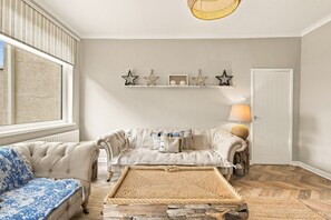 Un salon confortable avec un canapé, une table basse et une lampe. On aperçoit une fenêtre avec des stores et une porte au fond.