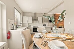A modern kitchen with white cabinets, a brick accent wall, and a dining area with a wooden table set for two. - Kilrenny - North East Escapes (Seahouses)