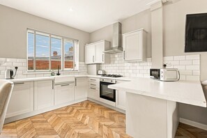 A modern kitchen with white cabinetry, a herringbone wood floor, and a window with a view of a brick building. - Kilrenny - North East Escapes (Seahouses)