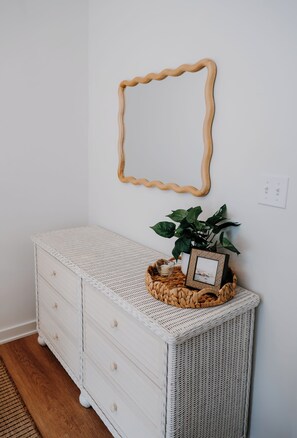 A white wicker dresser with drawers, a woven basket on top, a framed picture, and a green plant. - Bright and Cozy St. Augustine Home near Downtown (St. Augustine)