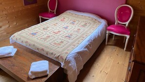 A cozy room with a wooden ceiling, a bed with a floral bedspread, two pink chairs, and a wooden table with towels. - Pink Room in a Wooden Chalet (Demi-Quartier)