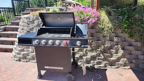 A portable grill with four control knobs and a digital display, placed on a brick patio with a stone wall and flowering plants in the background.