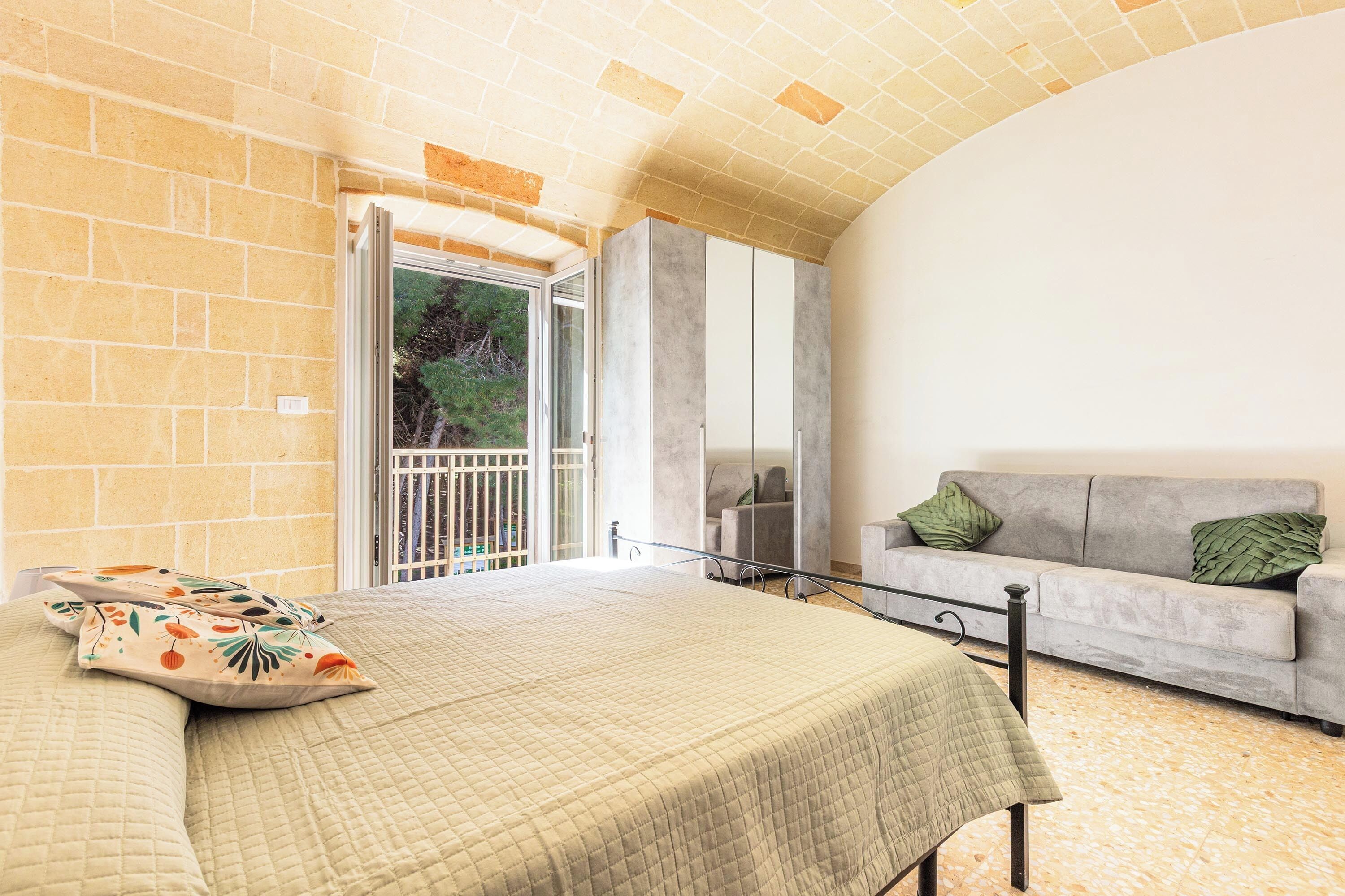 Ein Schlafzimmer mit Bett, einer Couch, einem Balkon mit Blick auf Bäume und einer Backsteinwand.