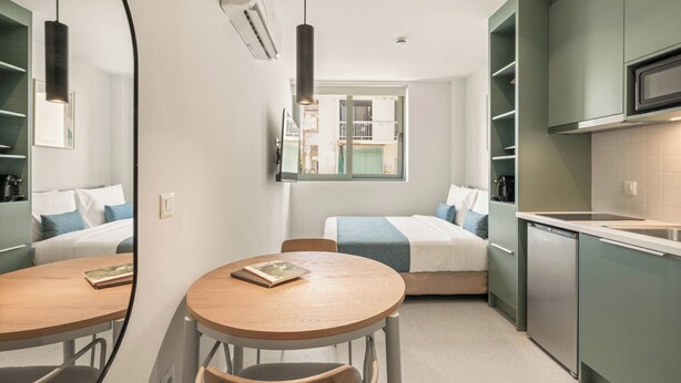 Una habitación de hotel pequeña con cama, una mesa redonda con sillas, una kitchenette y un espejo grande.