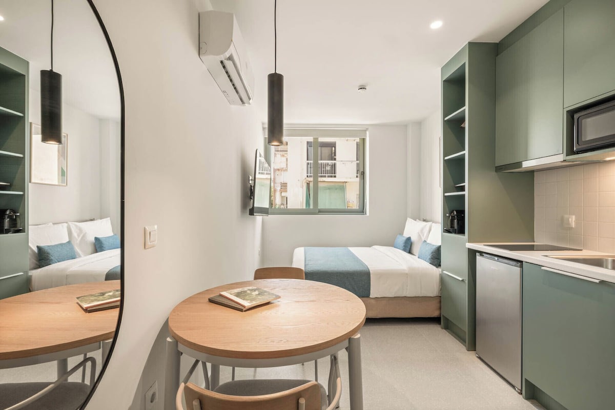 Quarto de hotel compacto com cama, mesa redonda com cadeiras, cozinha compacta e um espelho grande.