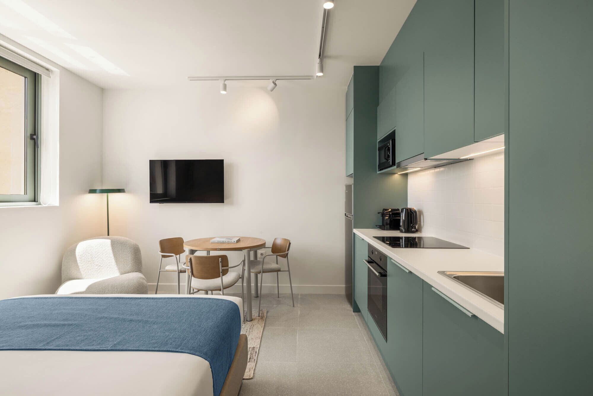 Ein kleines Hotelzimmer mit Bett, einer Sitzecke, einer Küchenzeile und einem an der Wand montierten Fernseher.