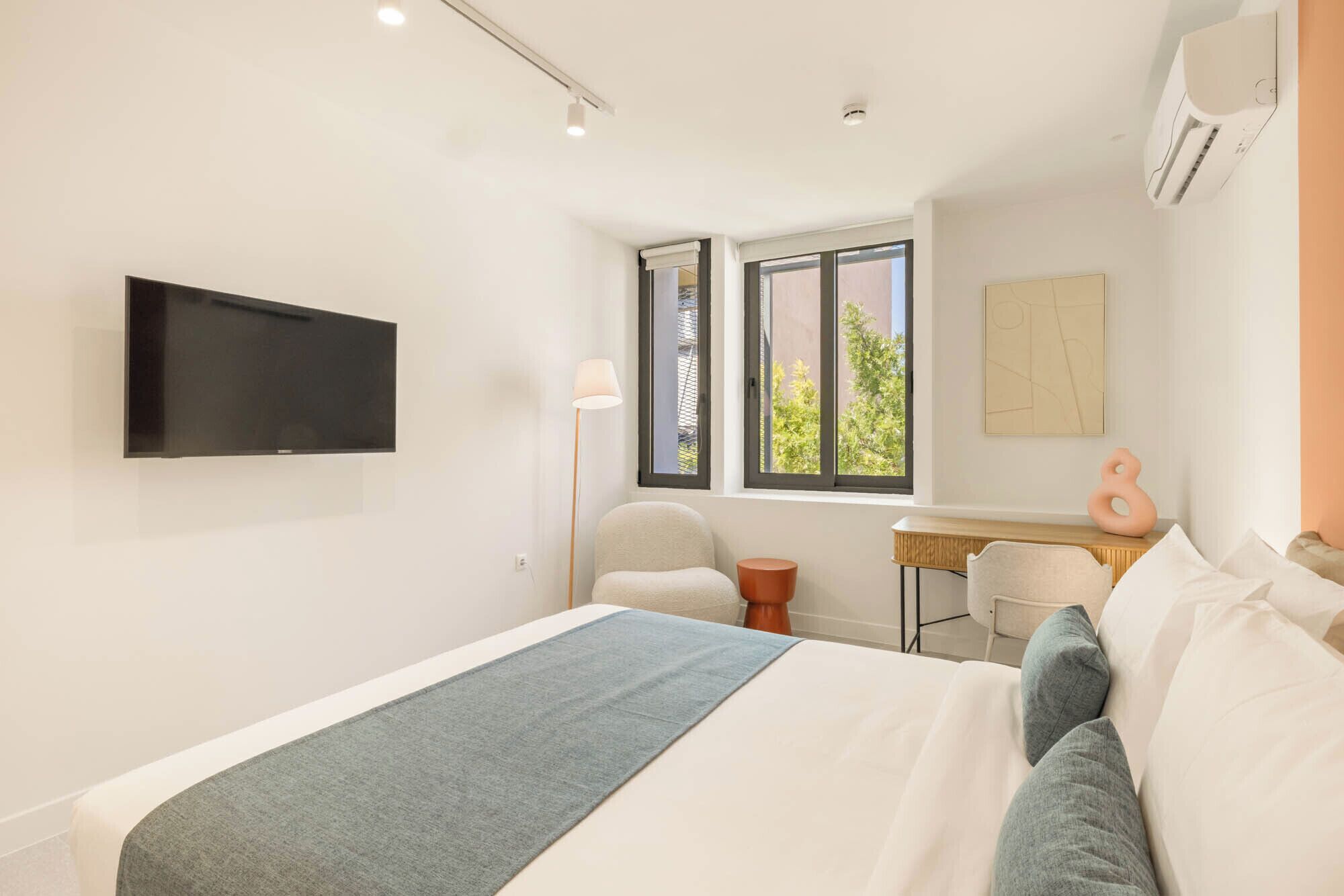 A hotel room with a bed, a desk, a chair, a TV mounted on the wall, and a window with a view of greenery.
