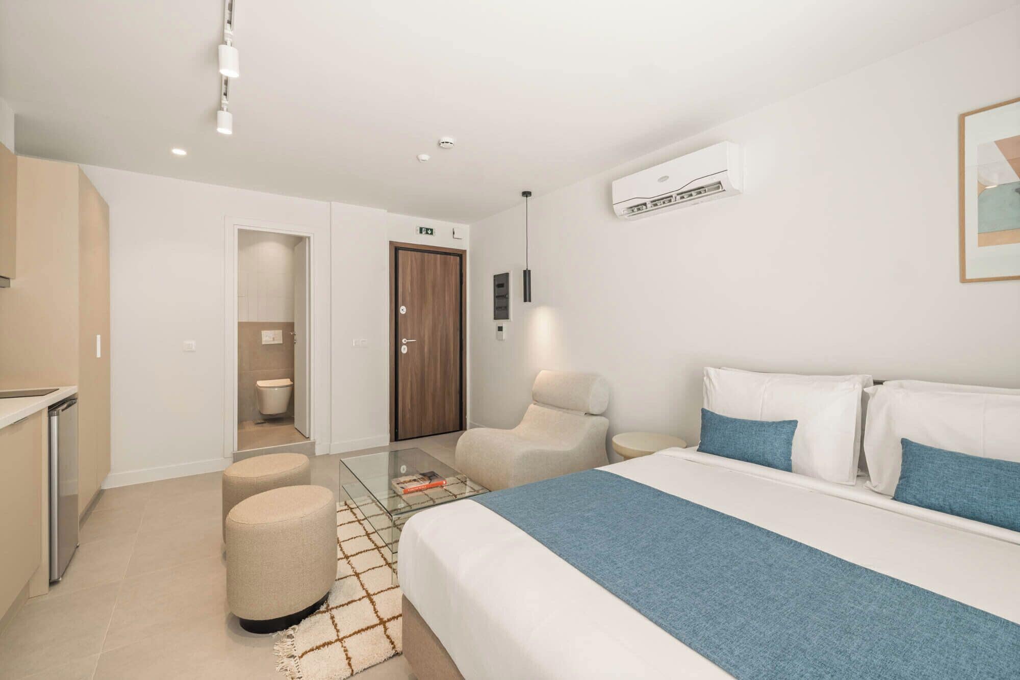 A hotel room with a bed, a chair, a small table, a bathroom, and an air conditioning unit.