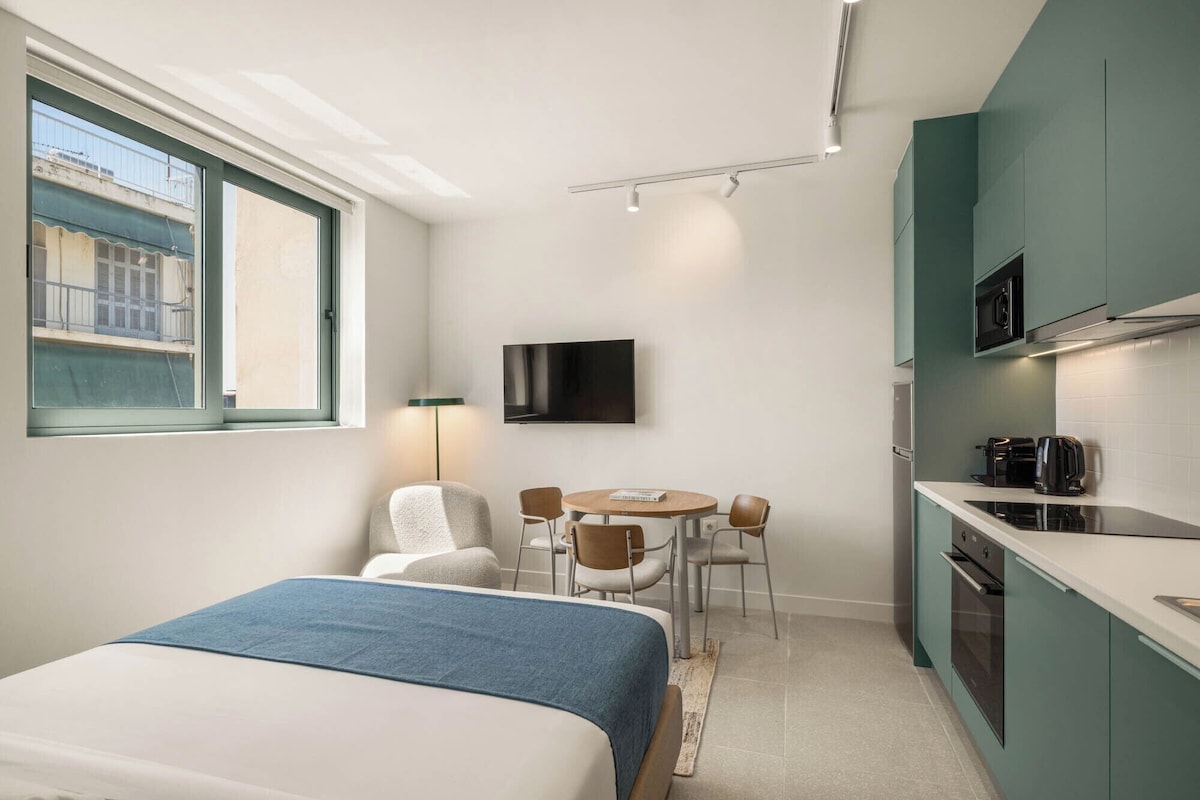 Quarto de hotel compacto com cama, pequeno espaço para refeições, cozinha compacta e TV na parede.