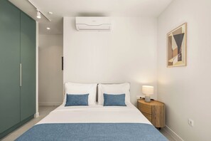 Hotellihuone, jossa on sänky, yöpöytä lampun kanssa, ilmastointilaite, vaatekaappi ja seinällä oleva kehystetty kuva.