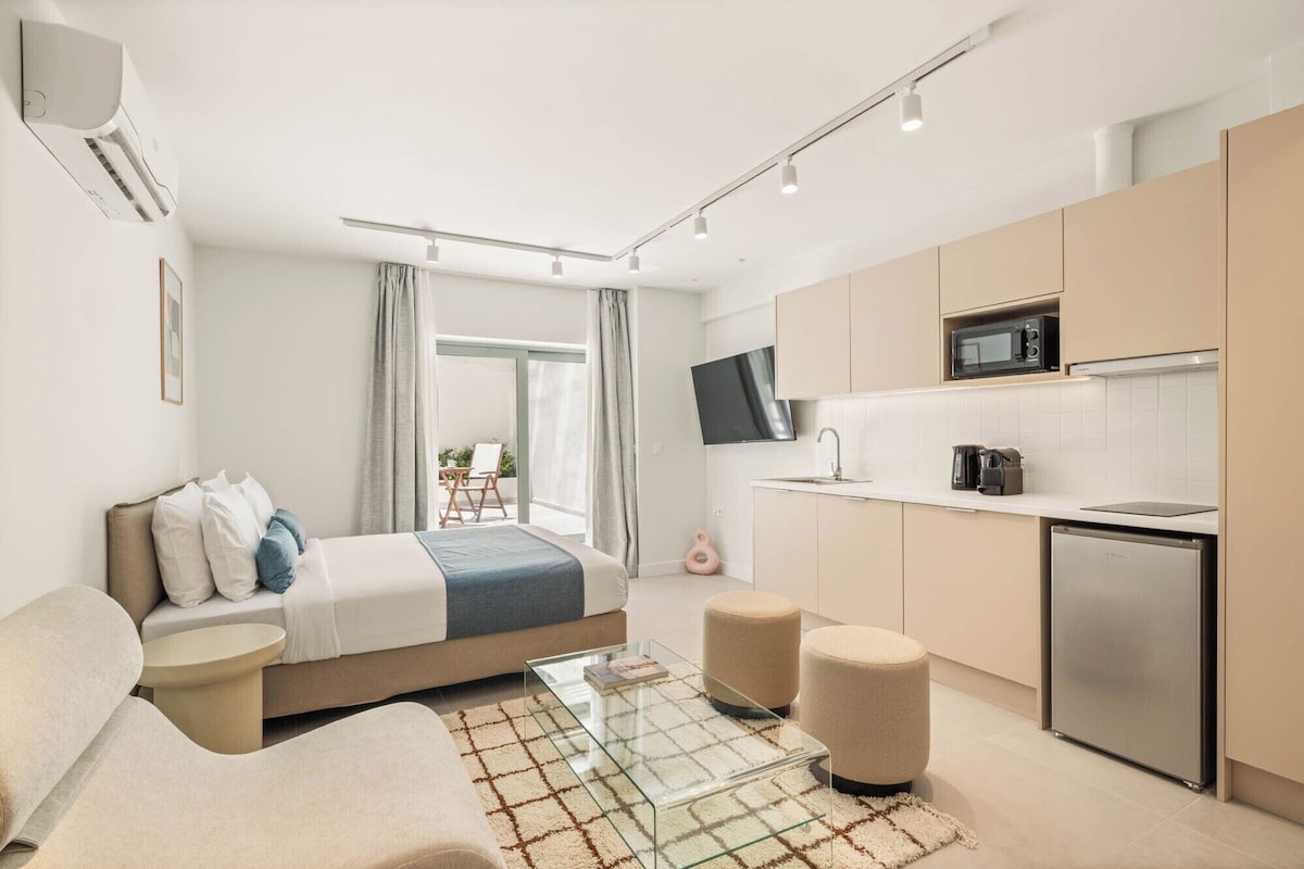 Quarto de hotel moderno com cama, mesa de centro de vidro, sofá, cozinha compacta e televisão.