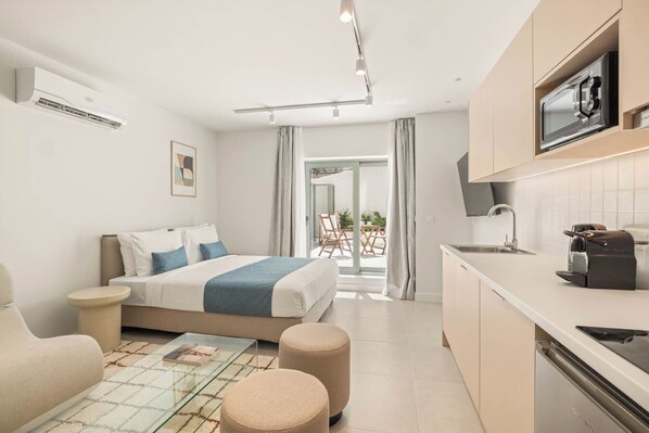 A modern hotel room with a bed, a small seating area, a kitchenette, and a view of the outdoors.