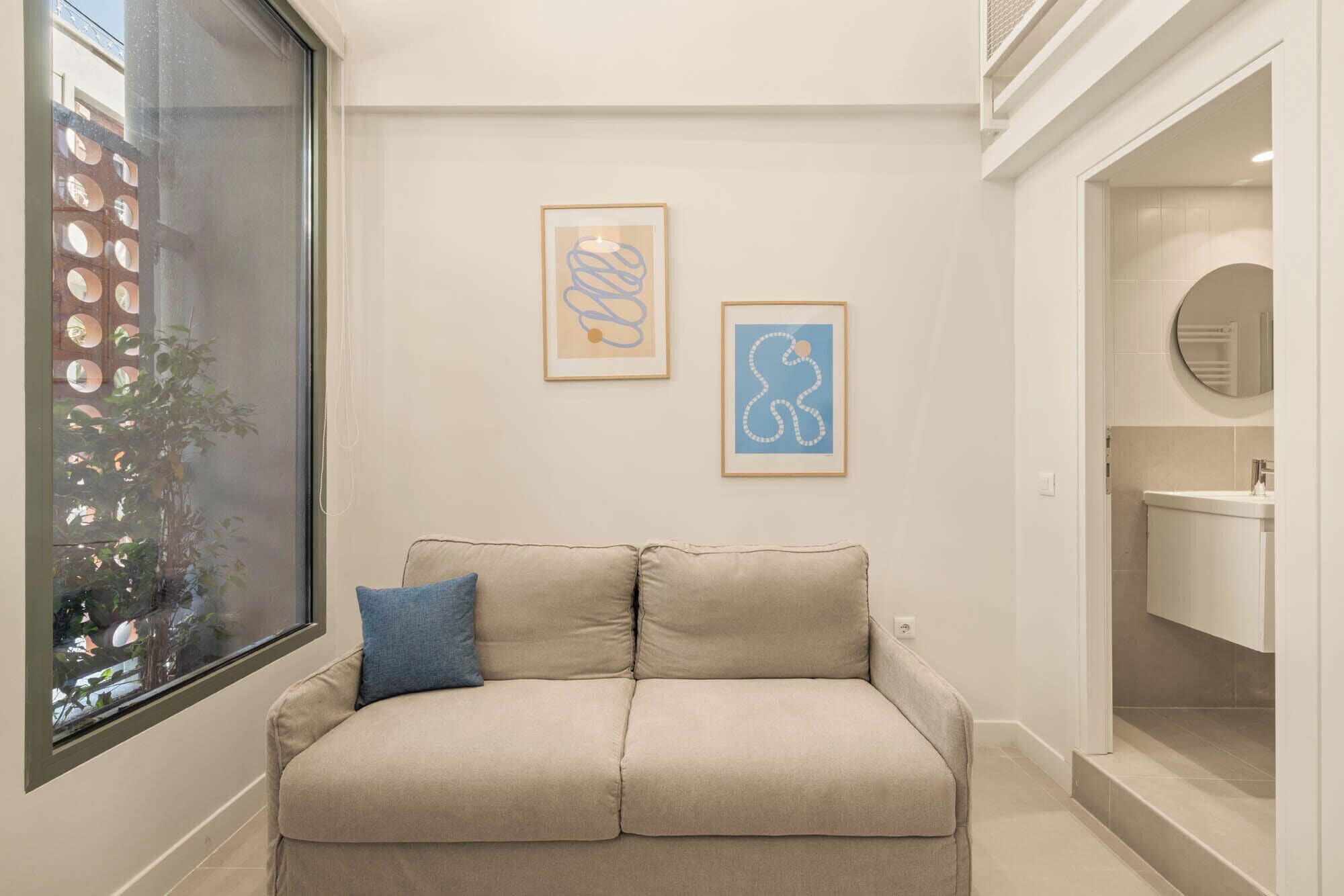 Una habitación en tonos neutros con un sofá color beige, dos obras de arte abstracto enmarcadas, un ventanal con vista a un edificio con ventanas circulares y un umbral que conduce a un baño.