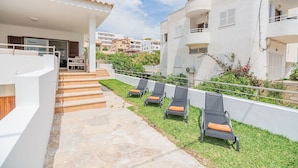 A residential building with a covered outdoor area, a staircase leading to a balcony, and a row of sun loungers on a grassy lawn. - Cosy Beach Apartment, Cala Sant Vicenc (Pollença)