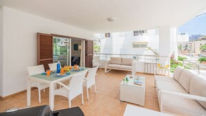 A balcony with a dining area, white wicker furniture, and a view of the city. - Cosy Beach Apartment, Cala Sant Vicenc (Pollença)