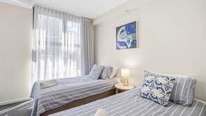 Ein Hotelzimmer mit zwei Betten, einem Fenster mit transparenten Vorhängen, einer Nachttischlampe und einem an der Wand hängenden abstrakten Gemälde.