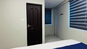 Una habitación con una puerta de madera oscura, una cama con colchón blanco, una ventana con persianas azules y suelo de baldosas.