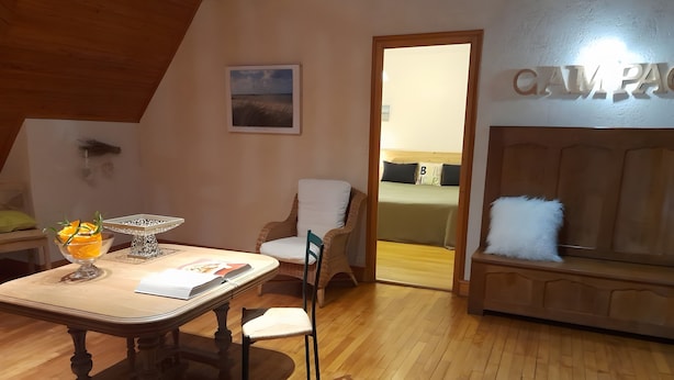 Une pièce avec un plancher en bois, une table à manger sur laquelle se trouve un saladier en verre rempli de fruits, une chaise, un canapé et un lit avec une pancarte indiquant « CAM. P. »