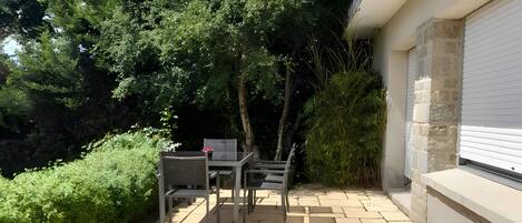 A paved patio area with a table and chairs, surrounded by greenery and a building with a stone facade.