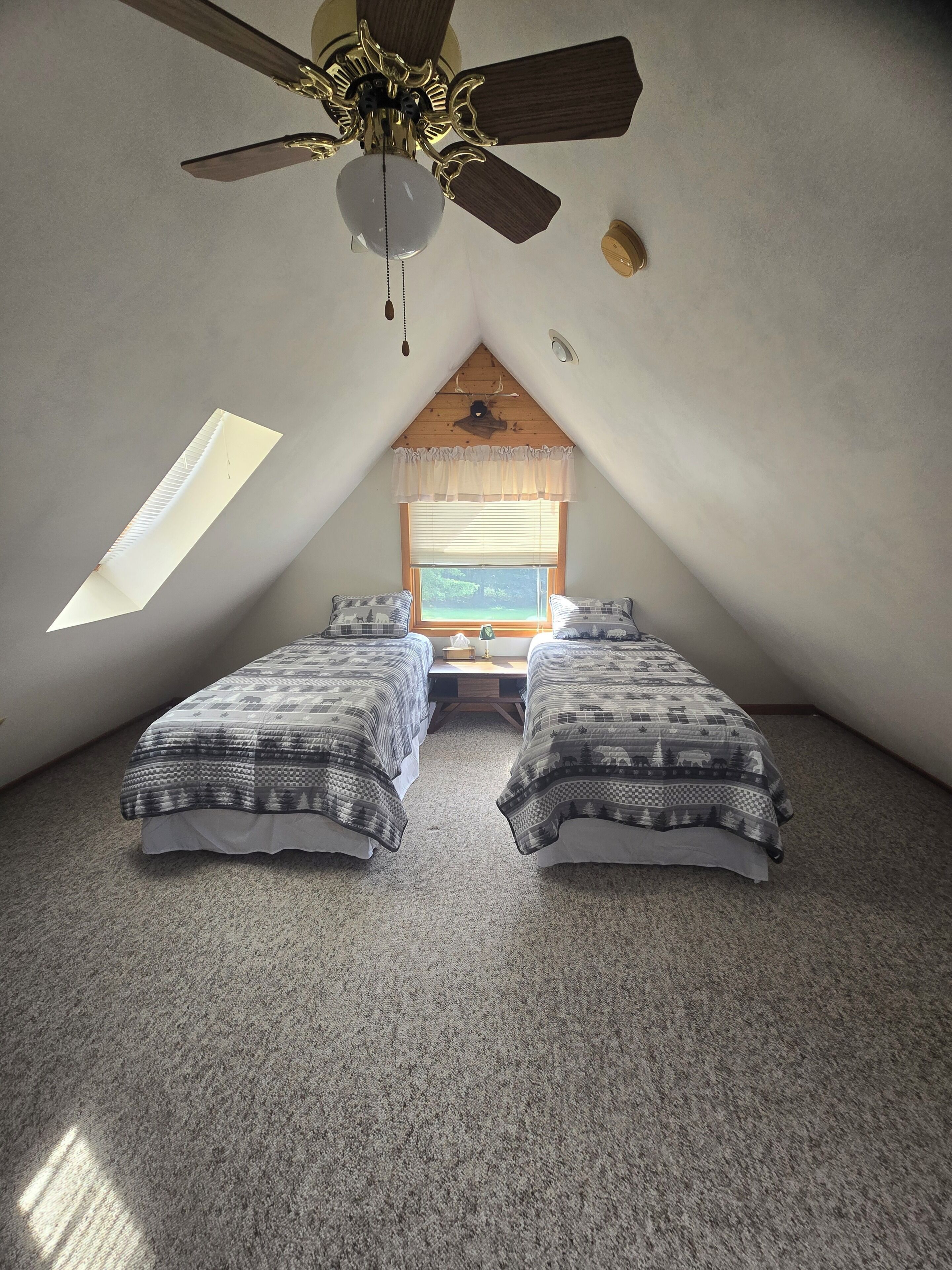 Ein kleines, gemütliches Schlafzimmer im Dachgeschoss mit zwei Betten, einem Deckenventilator, einem Fenster mit Jalousien und einer geneigten Decke.