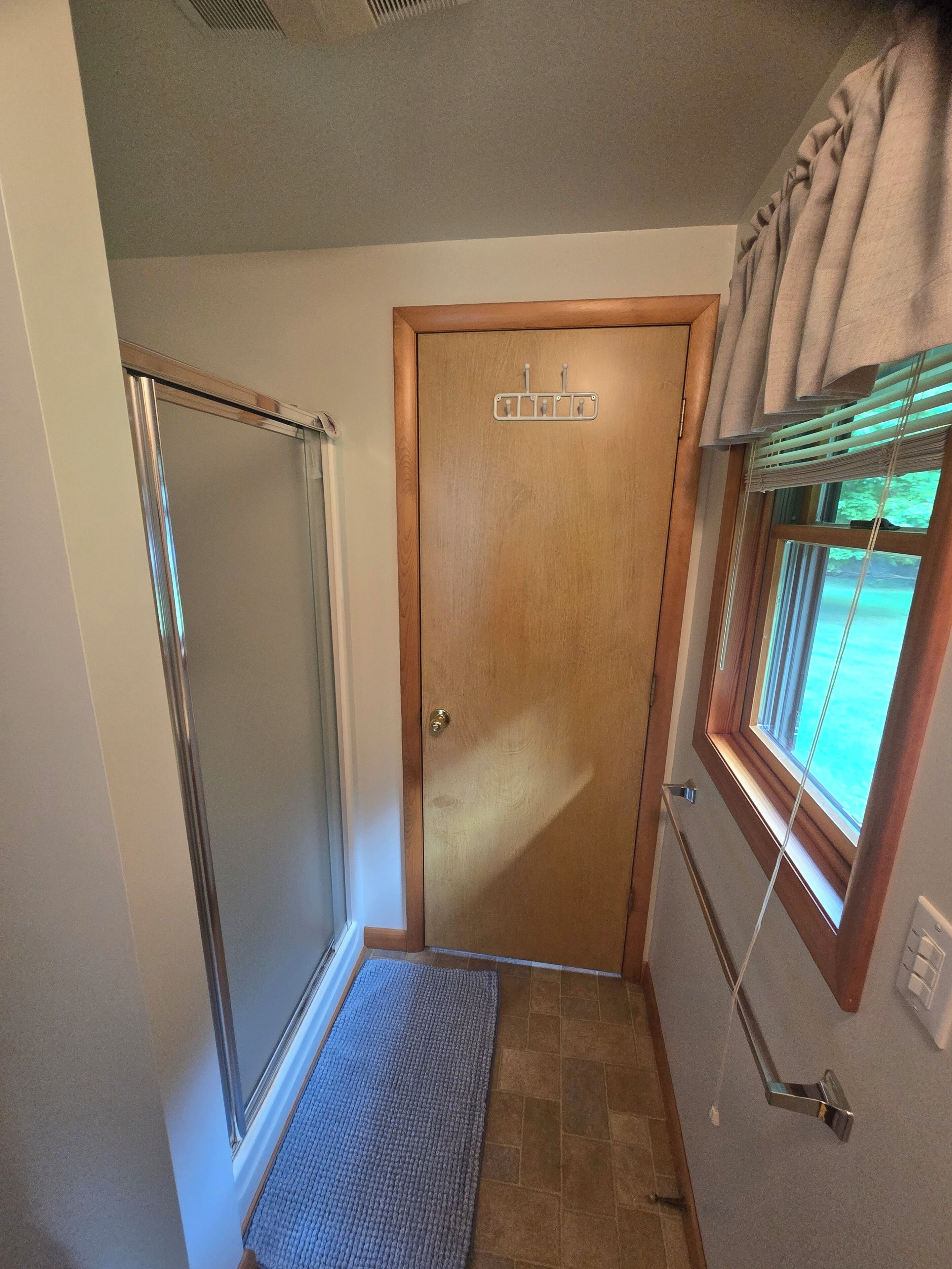 A bathroom with a shower, a door with a towel rack, and a window with blinds.
