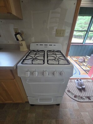 A kitchen with a white stove, wooden cabinets, and a window with a view of greenery. - Bluff Point Hideaway Private & Cozy 2 Bedroom Cabin (Keuka Park)