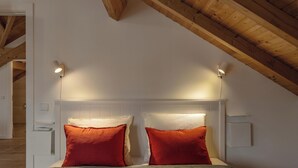 Un dormitorio con techo inclinado, cabecera blanca, dos almohadas rojas y lámparas de noche.