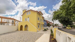 Un edificio amarillo con techo de tejas, un patio empedrado y una calle con coches aparcados.