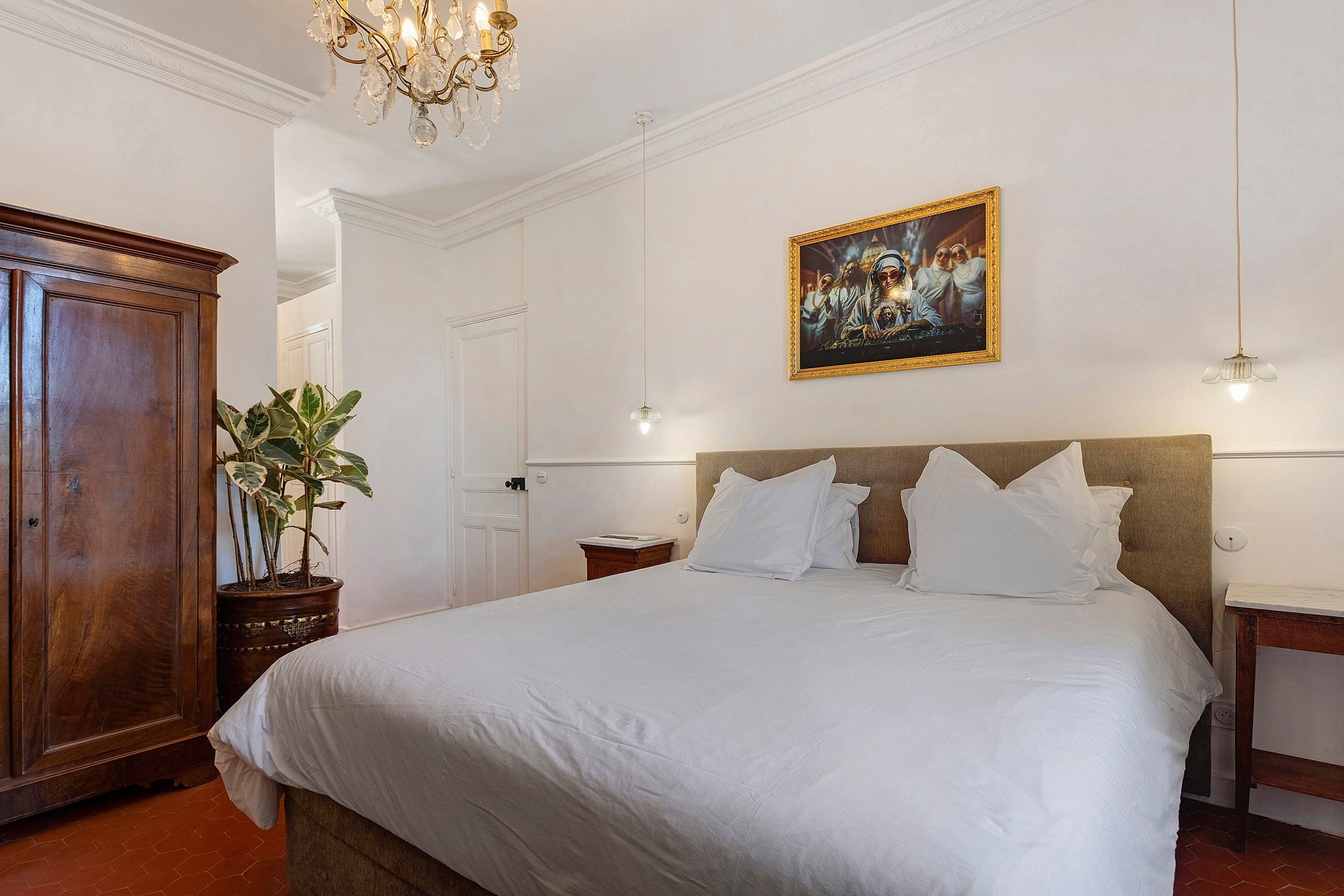 Una camera d'albergo con un letto grande, un armadio in legno, una pianta in vaso, un quadro appeso al muro e un lampadario.