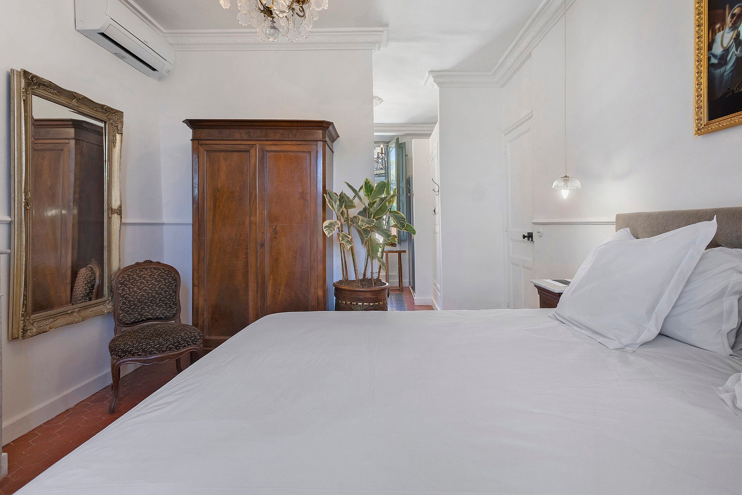 A bedroom with a large bed, a wooden wardrobe, a chair, a potted plant, and a mirror.