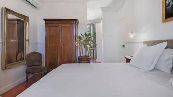 A bedroom with a large bed, a wooden wardrobe, a chair, a potted plant, and a mirror.