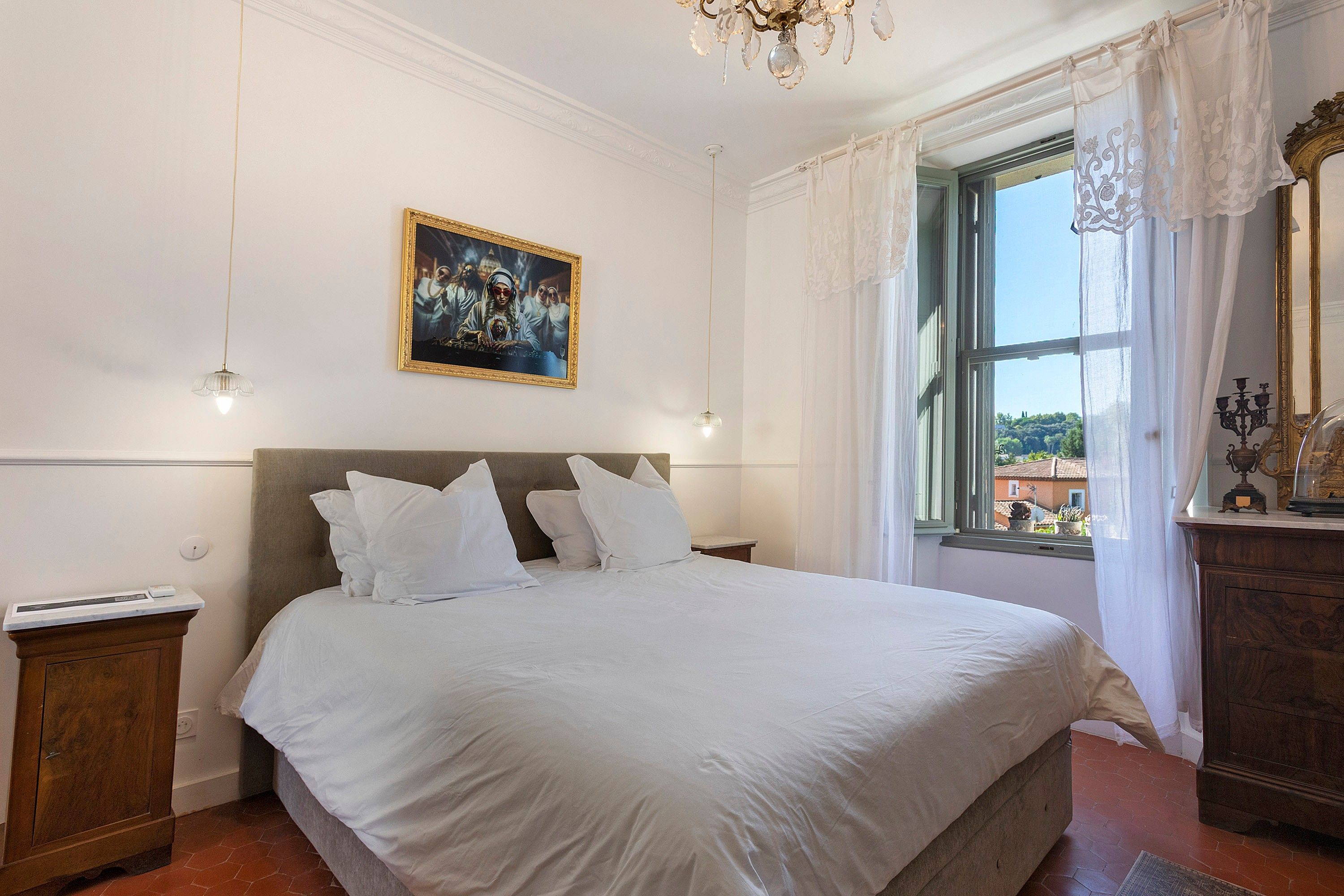 A bedroom with a large bed, a nightstand, a dresser, a window with curtains, and a chandelier.