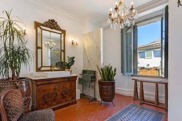 A well-lit room with a large mirror, a chandelier, a marble wall, and a view through a window.