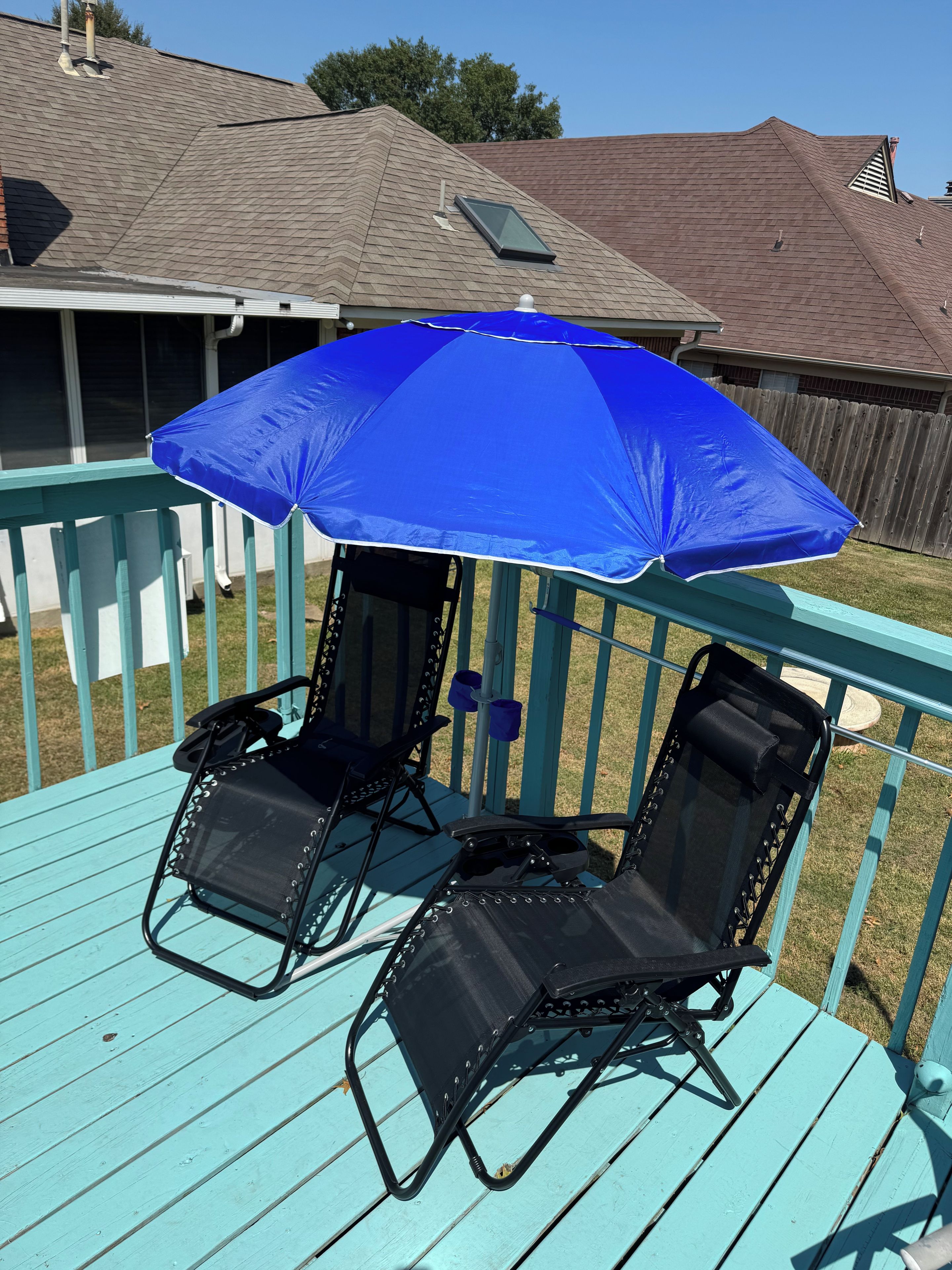 Un parasol bleu offre de l’ombre sur deux chaises longues noires, qui sont placées sur une terrasse turquoise.