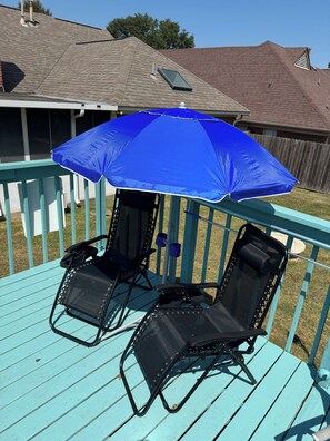 A blue umbrella providing shade over two black lounge chairs on a turquoise deck.