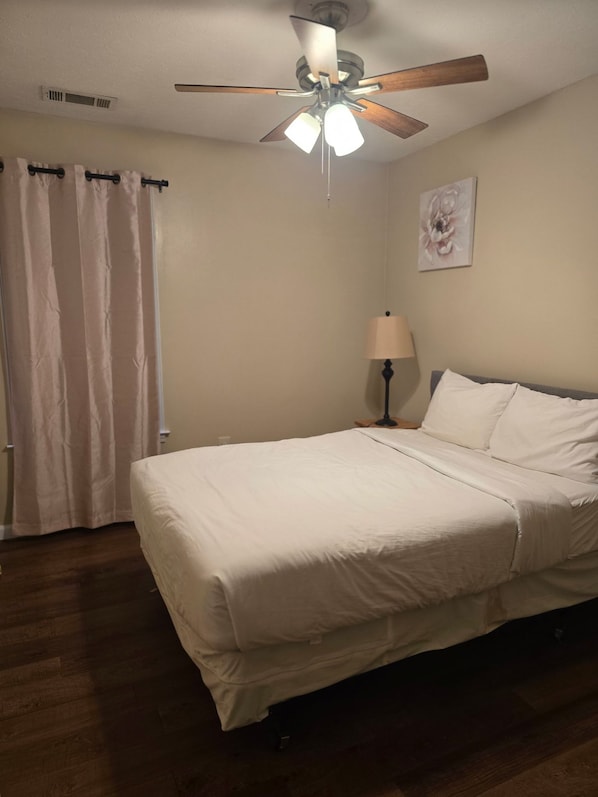 A hotel room with a bed, a ceiling fan, a nightstand with a lamp, and a window with curtains.