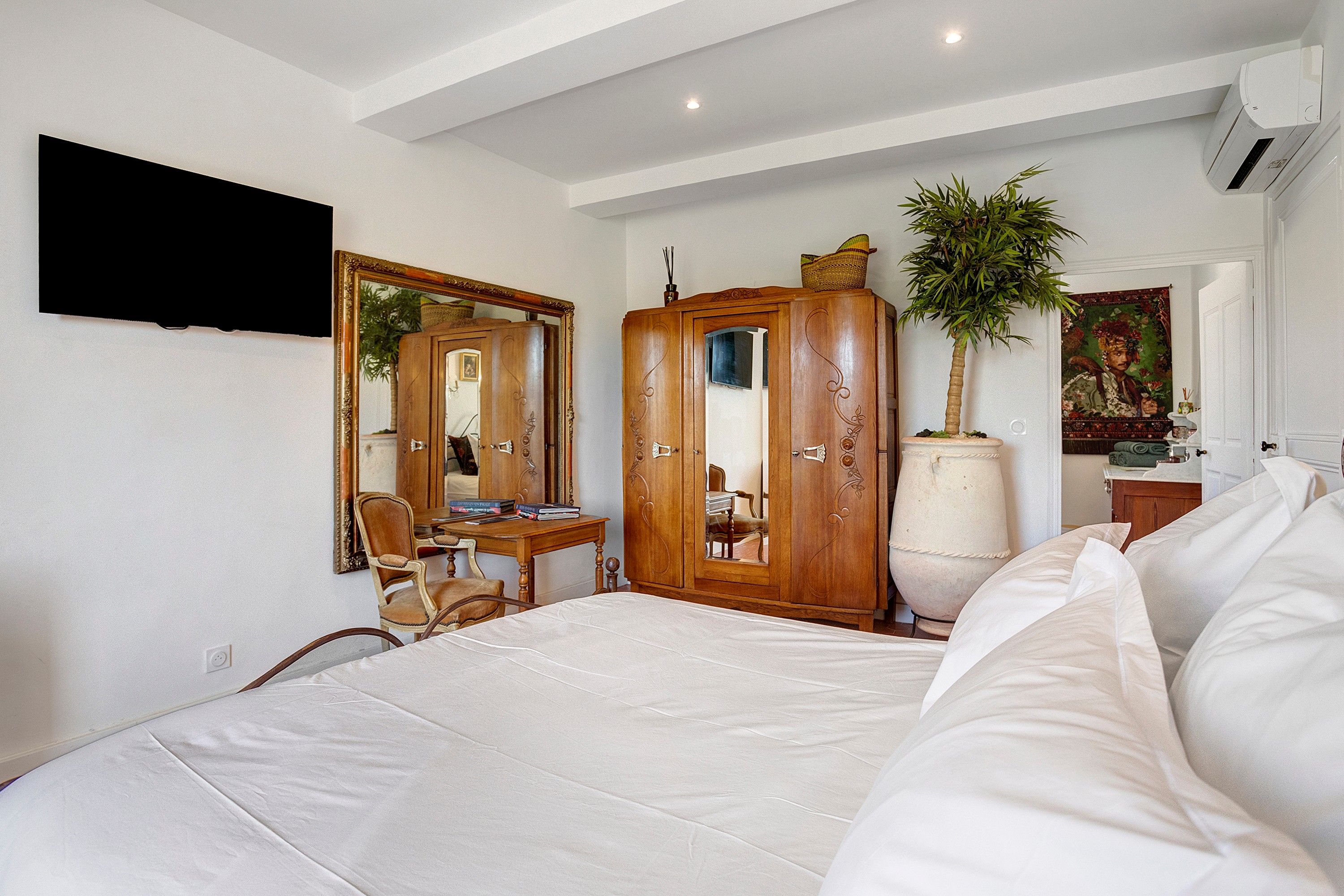 A bedroom with a bed, a wooden wardrobe, a desk with a chair, a large mirror, a potted plant, and a painting on the wall.