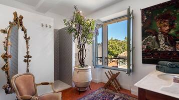 Une salle de bain avec un grand miroir, une plante en pot, une fenêtre donnant sur un beau paysage, un tableau et une chaise en bois.
