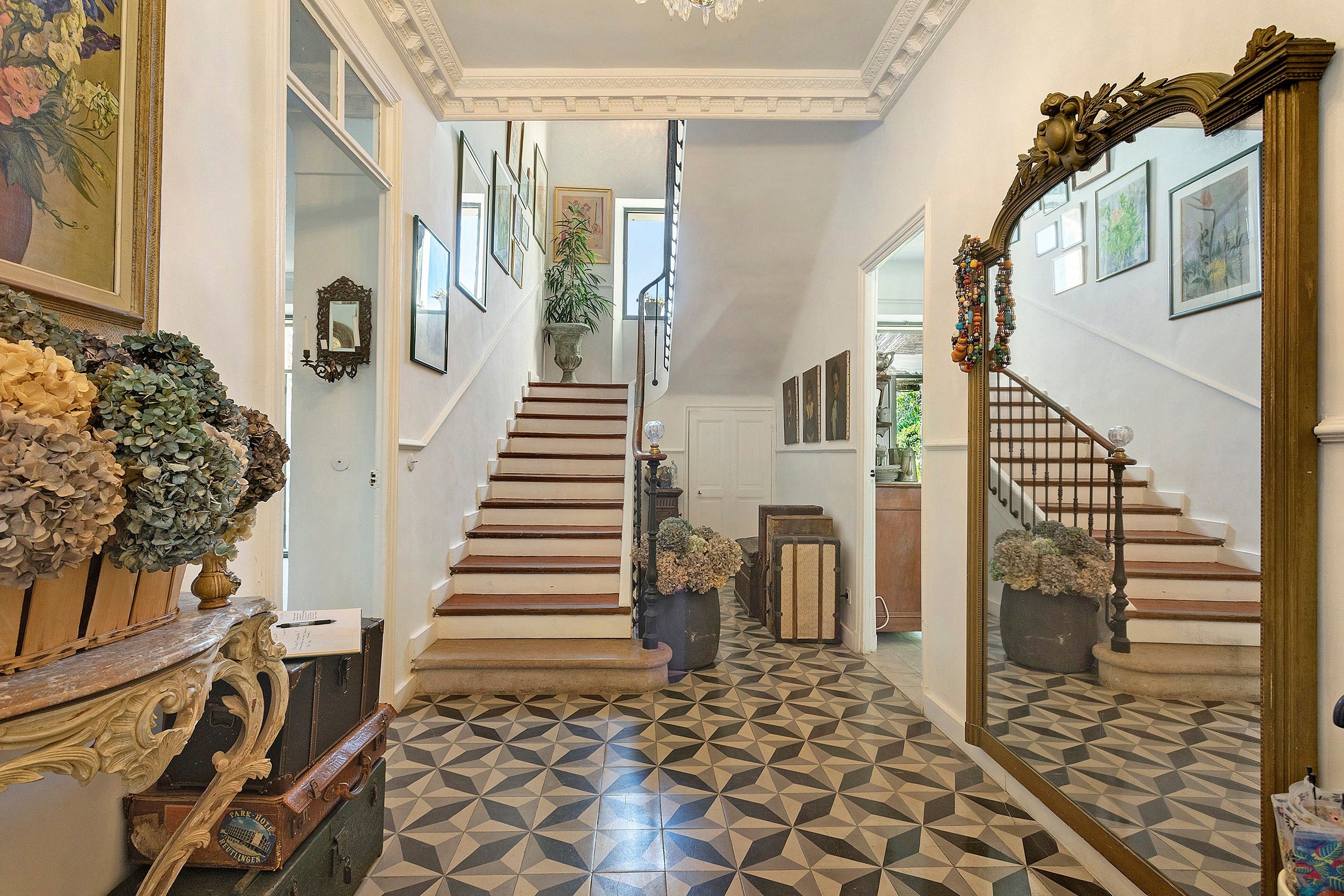 Un magnifique escalier, dont le sol est orné d’un motif géométrique, est bordé de tableaux encadrés et d’un miroir.