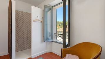 Ein Zimmer mit einem Fischgrätparkettboden, einem Holzstuhl mit einem gefalteten Handtuch, einer Tür mit einem hölzernen Kleiderbügel und einem Fenster mit Blick auf ein Ziegeldach und Grünflächen.