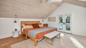 Ein Schlafzimmer mit schräg versiegelter Decke, einem Bett mit Holz Kopfteil, einer Bank, einem Nachttisch mit Pflanze und Blick ins Freie durch eine Glastür.
