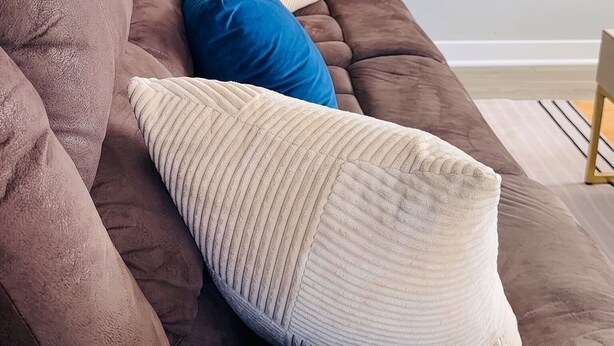 En brun sofa med en hvid og blå pude i et rum med en hvid dør og et vægmonteret fjernsyn.