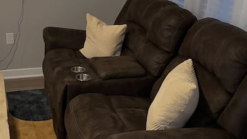 Un sofá de cuero marrón con dos cojines, un televisor de pantalla plana montado en la pared y una ventana con cortinas translúcidas.